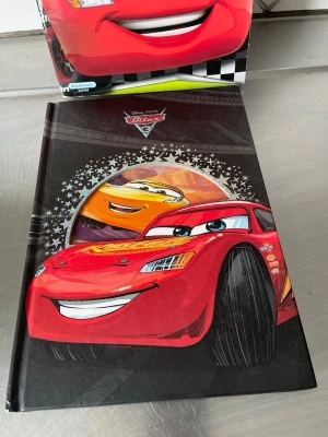 Disney Pixar Bilar bok+En bok med klistermärken  - En färgglad och cool bok från Disney Pixar Bilar med motiv av Blixten McQueen och Cruz Ramirez på hård pärm. Perfekt för dig som gillar fartfyllda äventyr och snygga illustrationer. Boken har svart bakgrund och tydliga, glada färger på omslagets. Medföljer en bok med delvis klistermärken i 