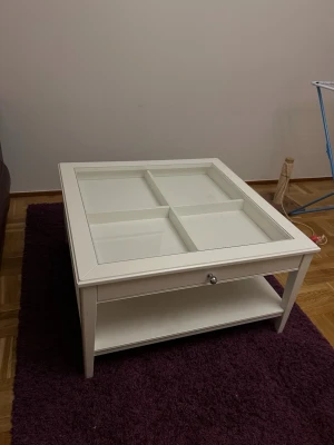 Vit soffbord med glasruta och låda - Säljer ett stilrent vitt soffbord i trä med fyrkantig form och glasskiva på toppen. Bordet har fyra fack under glaset, en praktisk låda med metallknopp och en extra hylla undertill för förvaring. Perfekt för att visa upp dekorationer eller förvara småprylar. Pickup only