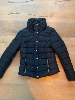 Svart pufferjacka med dragkedjor - Snygg svart pufferjacka med hög krage och silverfärgade dragkedjor på fickorna. Jackan har tryckknappar framtill och quiltad design som ger en cool look. Perfekt för kalla dagar och enkel att matcha med olika outfits.