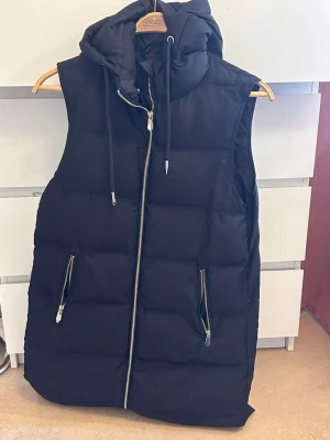 Svart lång dunväst med huva - Snygg svart dunväst med huva och dragkedja framtill. Västen har två fickor med dragkedjor och en quiltad design som ger en puffig look. Perfekt för lager på lager och streetstyle. Dragsko i huvan för extra stil och funktion.