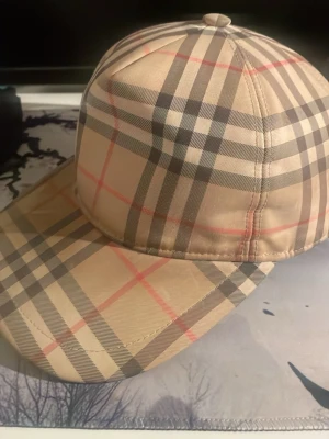Beige keps med rutigt mönster Burberry - Snygg beige keps från Burberry med det klassiska rutiga mönstret i svart, vitt och rött. Kepsen har böjd skärm och justerbar passform. Perfekt accessoar för en trendig och stilren look. Endast byten 