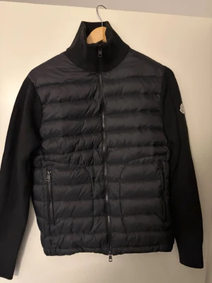 Moncler Cardigan  - Stilren svart pufferjacka från Moncler med quiltad front och stickade ärmar. Jackan har hög krage, två dragkedjefickor och Moncler-logga på ärmen. Använd ett par gånger annars i ett riktigt bra skick.