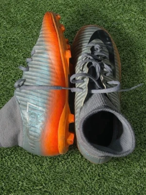 Nike Mercurial CR7 fotbollsskor - Snygga Nike Mercurial CR7 fotbollsskor med grå ovandel, svarta detaljer och orange sula. Skorna har snörning och en strumpliknande krage för extra stöd. Tillverkade i syntetmaterial som ger bra grepp och känsla på planen.