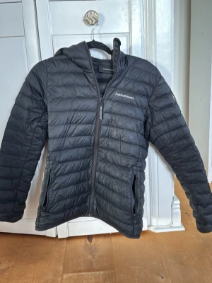 Svart dunjacka Peak Performance - Snygg svart dunjacka från Peak Performance med huva och dragkedja framtill. Jackan har två sidofickor med dragkedja och diskret logga på bröstet. 