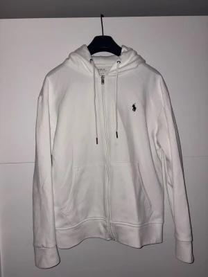 Vit hoodie från Polo Ralph Lauren - Säljer en stilren vit hoodie från Polo Ralph Lauren med dragkedja framtill, två fickor och justerbar huva med snören. Klassisk logga broderad på bröstet. Perfekt för en clean och avslappnad look. Tillverkad i mjukt bomullsmaterial. Skick 9/10