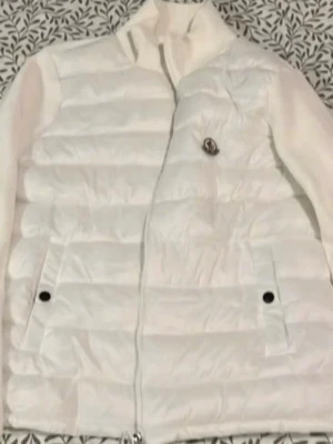 Vit cardigan från Moncler - Säljer en snygg vit dunväst från Moncler med klassisk quiltad design och hög krage. Västen har två fickor med tryckknappar och dragkedja framtill. Ikonisk Moncler-logga på bröstet och coolt serietidningsfoder på insidan.