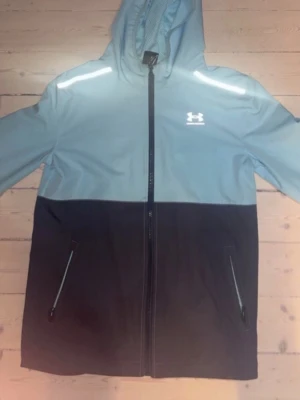 Under Armour tvåfärgad vindjacka - Snygg vindjacka från Under Armour i ljusblått och svart med hel dragkedja framtill. Jackan har huva, två sidofickor med dragkedja och reflekterande detaljer på axlarna. Perfekt för blåsiga dagar och sportiga outfits.