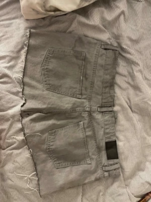 Grå denimshorts med råa kanter - Säljer ett par gråa denimshorts med råa, fransiga kanter och klassiska fem fickor. Shortsen har bälteshällor och silverfärgade knappar. Perfekta för en avslappnad och cool stil.
