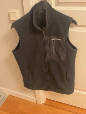Svart fleeceväst Peak Performance - Svart fleeceväst från Peak Performance med hög krage och hel dragkedja framtill. Västen har en bröstficka i nylon med logga samt två sidofickor med dragkedja. Perfekt lager för kyliga dagar och enkel att matcha till olika outfits. Det finns dock en pytte liten defekt på sista bilden. Men dragkedjan fungerar fortfarande som den ska.