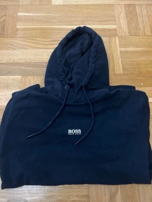 Svart hoodie från BOSS - Snygg svart hoodie från BOSS med klassisk logga på bröstet. Hoodien har huva med dragsko och en stor magficka framtill. Perfekt för en chill och stilren look.