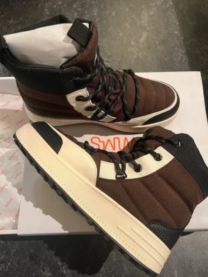 Oanvända SWIMS Snowjoggers 40 kängor - Ett par bruna vinterkängor från norska Swims. Perfekt för den som vill ha ett par bekväma och stilrena kängor. Unisex Stl 40. Nya i kartong  Normal i storlek Unisex-modell Vattentät ripstop-nylon Värmeisolerad  Snow Runner – tillverkad för både män och kvinnor – passar när din dagliga rutin kräver att du släpar dig igenom snöblandat regn, snöblandat regn eller snö. Ett praktiskt val för vintern, det vattentäta membranet inuti håller fötterna torra, och den är tillverkad med en tjock sula.