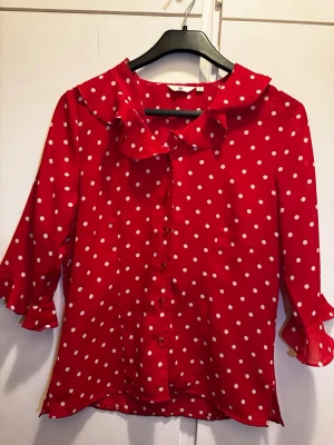 Röd prickig blus med volangkrage - Röd blus med vita prickar och volangkrage. Blusen har knappar framtill och trekvartsärm med volangdetaljer vid kragen och ärmslut. Ett lätt och luftigt material som ger en skön känsla. Perfekt för dig som gillar retroinspirerad stil.🌸