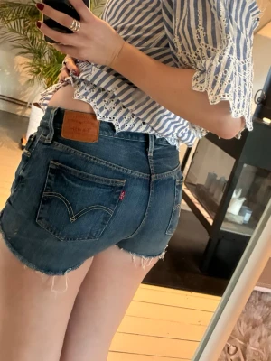 Levi's jeansshorts i mörkblå denim - Snygga mörkblå jeansshorts från Levi's med råa kanter och klassiska bakfickor. Modellen är lågmidjad och har en avslappnad passform. Perfekta till sommaren och lätta att matcha med olika toppar.