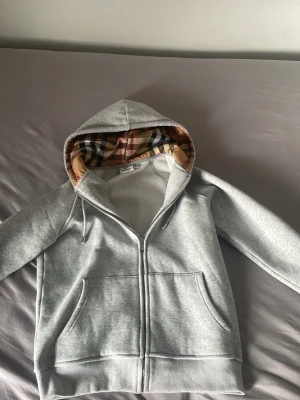 Grå zip hoodie från Burberry - Säljer en grå zip hoodie från Burberry med klassiskt rutigt foder i huvan. Tröjan har dragkedja framtill, två fickor och justerbar huva sopn ger en clean och stilren look. Perfekt för dig som gillar exklusiva streetwear-plagg. Dessutom kan jag diskutera pris!