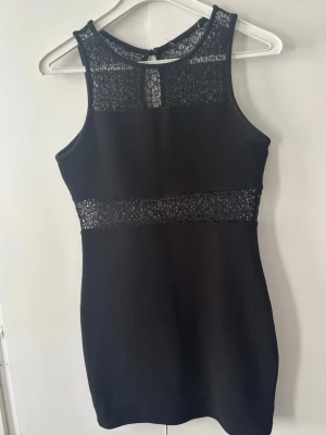 Svart klänning med spetsdetaljer - Snygg svart kortklänning med spetsdetaljer upptill och i midjan. Klänningen är ärmlös och har en rund halsringning med transparent spets som ger en elegant touch. Perfekt för dig som vill ha en stilren och modern look.