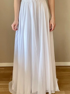 Vit långklänning med spetsdetaljer - Elegant vit bröllopsklänning med vackra spetsdetaljer upptill och ett svepande, luftigt fall. Klänningen har en  liten fläck på kupan inne i klänningen. Men absolut inget som syns! 