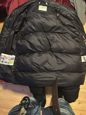 Svart dunjacka från Moncler - Snygg svart dunjacka från Moncler med stora, vadderade paneler och dragkedja framtill. Jackan har huva med Moncler-logga längs kanten och praktiska fickor med dragkedja. Perfekt för kalla vinterdagar och riktigt trendig stil.