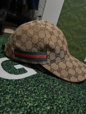 Gucci beige GG keps - Säljer en beige keps från Gucci med det klassiska GG-monogrammet över hela kepsen. På sidan finns en grön och röd rand som signaturdetalj. Kepsen har böjd skärm och justerbar rem bak. 