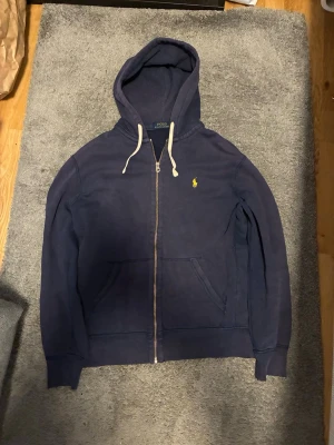  Polo Ralph Lauren Zip Hoodie - Snygg marinblå hoodie från Polo Ralph Lauren med dragkedja framtill, vita dragsnören och klassisk gul logga på bröstet. Hoodien har kängurufickor och ribbade muddar vid ärmar och nederkant. Perfekt för en avslappnad och stilren look.
