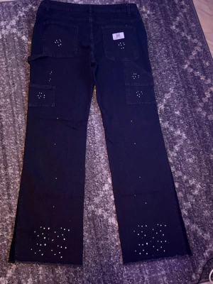 Svarta cargojeans med strassdetaljer - Säljer ett par svarta cargojeans med raka ben och flera fickor. Jeansen har coola strassdetaljer på fickor och nedtill på benen som ger en unik look. Perfekta för dig som vill sticka ut och gillar streetstyle vibes. Materialet är klassisk denim.