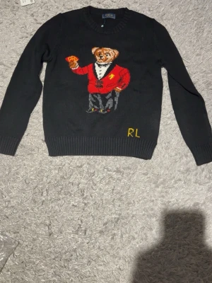 Svart stickad tröja med björnmotiv RL - Stickad svart tröja från Polo Ralph Lauren med ikoniskt björnmotiv på bröstet. Björnen har röd kavaj och håller spelkort. RL-broderi i gult nedtill. Tröjan har rund hals och ribbade muddar. Perfekt för dig som gillar statement-plagg.