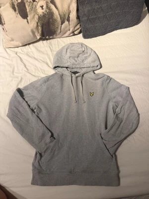 Grå Lyle & Scott hoodie - Snygg grå hoodie från Lyle & Scott med klassisk huva och dragsnören. På bröstet sitter den ikoniska gula örnloggan broderad. Hoodien är tillverkad i mjukt bomullsmaterial och har ribbade muddar vid ärmslut och nederkant. Perfekt för en chill och stilren look. Inga skador eller slitningar på den, använt i bra skick! 