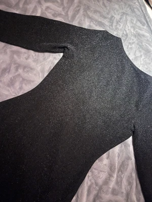 Svart glittrig långärmad klänning - Snygg svart klänning med glittrigt tyg och lång ärm. Klänningen har en kroppsnära passform och är perfekt för dig som vill sticka ut. Materialet känns mjukt och stretchigt, vilket gör den bekväm att bära hela kvällen.