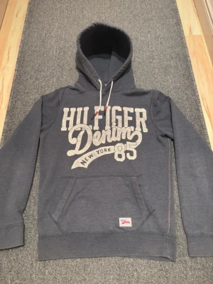 Grå Hilfiger Denim hoodie - Snygg grå hoodie från Hilfiger Denim med stor vit logga och text på bröstet. Klassisk känguruficka framtill, dragsko i huvan och mjukt material. Perfekt för en avslappnad streetstyle-look.