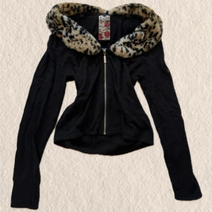 Svart croppad ziptröja med fuskpäls - Säljer en svart croppad ziptröja med lång ärm och dragkedja framtill. Tröjan har en stor krage i leopardmönstrad fuskpäls som ger en edgy vibe. Perfekt för dig som gillar Y2K-stil och vill sticka ut lite extra.