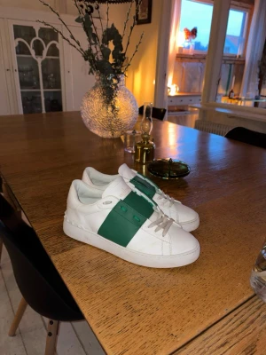 Valentino sneakers med grön detalj - Snygga vita sneakers från Valentino med en bred grön remsa över sidan och klassisk snörning. Skorna har rund tå och platt sula, tillverkade i skinn för en exklusiv känsla. Perfekta för dig som vill ha en stilren men ändå unik sneaker.