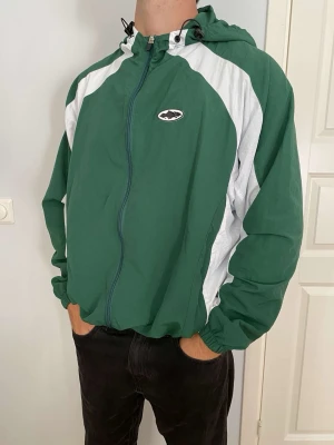 Grön och vit Corteiz jacka  - Snygg grön vindjacka från Corteiz med vita detaljer längs ärmar och axlar. Jackan har huva med dragsko, dragkedja framtill och elastiska muddar vid ärmslut och midja. Liten logga på bröstet och broderad text på ryggen. Perfekt för vår eller höst. 