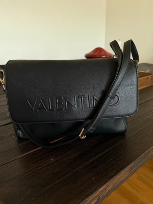 Svart axelväska från Valentino - Snygg svart axelväska från Valentino i slätt konstläder med präglad logga på framsidan. Väskan har justerbar axelrem och guldfärgade metalldetaljer med Valentino-gravering. Perfekt storlek för det viktigaste och stilren design som passar till många outfits.