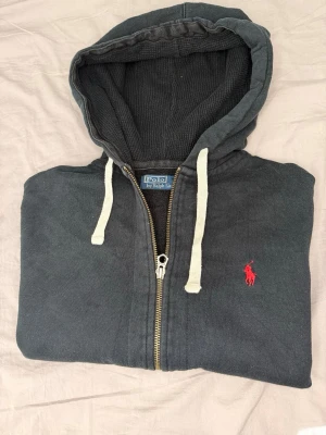Mörkgrå hoodie från Polo Ralph Lauren - Snygg mörkgrå hoodie med dragkedja från Polo Ralph Lauren. Klassisk design med vit dragsko i huvan och röd broderad logga på bröstet. Tillverkad i mjuk bomull, perfekt för en avslappnad stil. Praktiska fickor framtill och ribbade muddar.