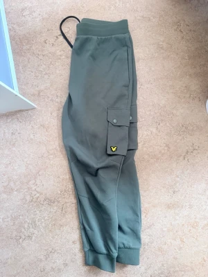 Gröna cargobyxor från Lyle & Scott size XL - Snygga gröna cargobyxor från Lyle & Scott med resår i midjan och svarta dragsnören. Byxorna har en stor ficka på benet med knapp och den klassiska gula loggan. Tillverkade i mjukt bomullsmaterial och har muddar vid bensluten för en avslappnad look.