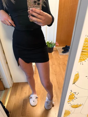 Svart kortkjol med slits - Säljer en svart kortkjol med slits på sidan. Kjolen har en tight passform och är enkel i designen, vilket gör den lätt att matcha med olika toppar. Perfekt för dig som gillar en stilren och trendig look.