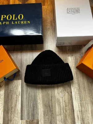 Svart mössa Acne Studios - Säljer en svart ribbstickad mössa från Acne Studios med uppvikt kant och den klassiska Acne-loggan framtill. Mössan är gjord i mjukt material och har en enkel, stilren design som passar till det mesta.