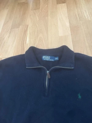 Mörkblå stickad tröja Polo Ralph Lauren - Mörkblå stickad tröja från Polo Ralph Lauren med halv dragkedja och ribbad krage. Tröjan har det klassiska gröna Polo-märket broderat på bröstet och är tillverkad i ett mjukt stickat material. Perfekt för en chill och stilren look.