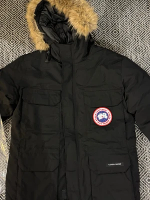 Svart parkas från Canada Goose - Säljer en svart parkas från Canada Goose med ikonisk röd och vit logotyp på bröstet. Jackan har stor huva med beige fuskpäls, flera praktiska fickor och stängs med dragkedja och vindslå. Perfekt för kalla dagar och riktigt snygg streetstil. 