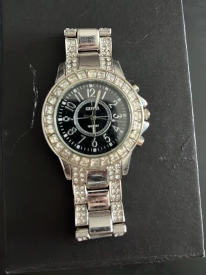 Silverfärgad klocka från Guess - Stilren klocka från Guess i silverfärg med svart urtavla och tydliga vita siffror. Armbandet och boetten är dekorerade med gnistrande stenar som ger en lyxig känsla. Perfekt accessoar för dig som vill sticka ut lite extra. (Den kommer inte med lådan)