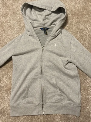 Grå hoodie från Polo Ralph Lauren - Snygg grå hoodie med dragkedja från Polo Ralph Lauren. Tröjan har huva, fickor framtill och den klassiska lilla loggan broderad på bröstet. Perfekt för en avslappnad och stilren look.