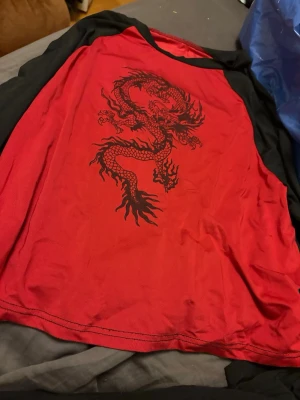 Röd och svart t-shirt med draktryck - Cool röd t-shirt med svarta ärmar och ett stort svart drakmotiv på framsidan. Tröjan har rund halsringning och är tillverkad i ett mjukt material som känns skönt mot huden. Perfekt för dig som vill sticka ut med en unik design.