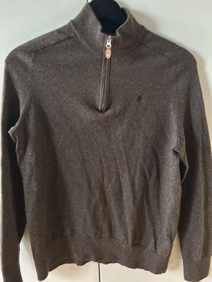 Mörkbrun stickad tröja Morris - Mörkbrun stickad tröja från Morris med halv dragkedja och hög krage. Tröjan har ett diskret broderat Morris-emblem på bröstet och ribbade muddar vid ärmslut och nederkant. Perfekt för lager på lager och snygg till jeans.