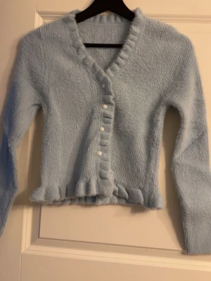 Ljusblå fluffig kofta med volang - Supermjuk ljusblå kofta med volangkant och pärlknappar. Koftan har lång ärm, v-ringning och är gjord i ett fluffigt material som känns riktigt mysigt. Perfekt för lager-på-lager och ger en söt touch till din outfit.