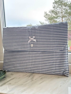 Blårandig datorväska med rosett - Snygg datorväska i blått och vitt med horisontella ränder. Väskan har en vadderad design, stängs med tryckknapp och är dekorerad med en liten vit rosett på framsidan. Perfekt för dig som vill ha en söt och stilren look till din laptop för jobb, skola eller vardag!💗🎀