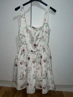 Vit blommig kortklänning - Söt vit kortklänning med breda axelband och blommigt mönster i rosa, grönt och vinrött. Klänningen har en utställd kjol och markerad midja, perfekt för varma dagar. Den är i storlek 34