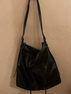 Svart axelväska från Zara - Snygg svart axelväska från Zara i mjukt konstläder. Väskan har en slouchy form, justerbart axelband och dragkedja på insidan. Perfekt för dig som gillar minimalistisk och stilren design.