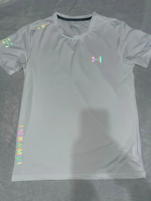 Vit tränings-tshirt från Under Armour - Snygg vit tränings-tshirt från Under Armour med regnbågsfärgade loggor och detaljer. Tillverkad i ett lätt och snabbtorkande material som passar perfekt till gymmet eller löprundan. Rund halsringning och korta ärmar för maximal rörelsefrihet.