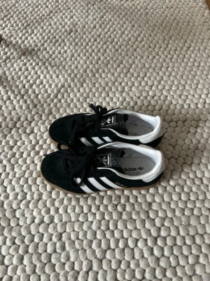 Adidas Gazelle svarta sneakers - Svarta Adidas Gazelle sneakers med klassiska vita ränder och vit häl. Skorna har ovandel i mocka, snörning och gummisula i brunt. 