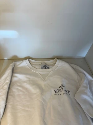 Vit sweatshirt från Replay - Säljer en stilren vit sweatshirt från Replay med trycket 'Replay Blue Jeans' på bröstet. Tröjan har rund halsringning och klassisk passform, perfekt för en avslappnad look. Materialet känns mjukt och bekvämt mot huden.