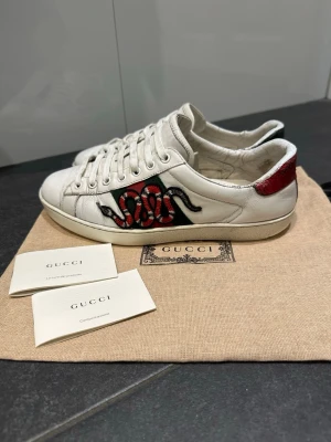 Gucci Ace sneakers med orm-brodyr - Tja säljer nu mina Gucci Ace sneakers i vitt skinn med ikonisk orm-brodyr.Baksidan har metallicdetaljer i rött och grönt. Klassisk låg modell med snörning och rund tå. Snygg streetstil med exklusiv känsla. Kan även byta mot en Gucci keps !